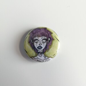 Puede incluir: Un bot&oacute;n redondo con una ilustraci&oacute;n estilizada de una persona con cabello morado, maquillaje oscuro y un collar negro. El fondo es amarillo p&aacute;lido con acentos verdes. La obra de arte tiene una est&eacute;tica g&oacute;tica o alternativa.