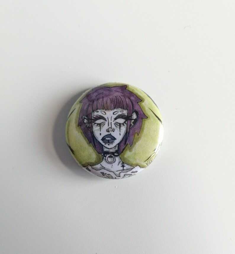 Puede incluir: Un bot&oacute;n redondo con una ilustraci&oacute;n estilizada de una persona con cabello morado, maquillaje oscuro y un collar negro. El fondo es amarillo p&aacute;lido con acentos verdes. La obra de arte tiene una est&eacute;tica g&oacute;tica o alternativa.