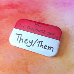 May include: A red and white button with the text "Hi! My Pronouns are... They/Them" written in black marker.