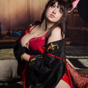 Peut inclure: Une femme portant un kimono noir et rouge avec des broderies dorées, des oreilles de chat roses et un soutien-gorge en dentelle rouge. Elle est assise sur un sol avec un fond en bois.