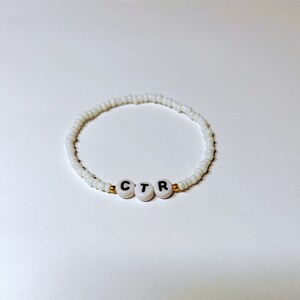 Op de afbeelding: Wit kralenarmband met gouden accenten en de letters "CTR" in zwart op witte kralen.