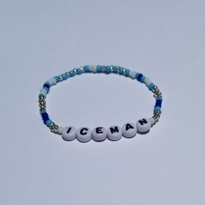 May include: A white beaded bracelet with blue and silver beads. The bracelet spells out the word "ICEMAN" in black letters on white beads.