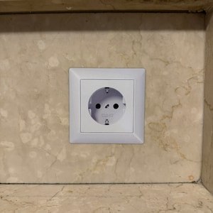 Könnte beinhalten: Eine weiße Steckdose mit zwei runden Löchern und einem Erdungsstift ist in eine hellbeige Marmorwand eingelassen. Die Steckdose hat einen quadratischen weißen Rahmen.