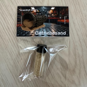 Könnte beinhalten: Ein kleines Glasfläschchen mit schwarzem Deckel, gefüllt mit einer hellbraunen, körnigen Substanz, ist in einem durchsichtigen Plastikbeutel versiegelt. Eine Karte hinter dem Beutel zeigt eine mechanische Illustration und den Text "GrainShift 9000" und "Getriebesand".