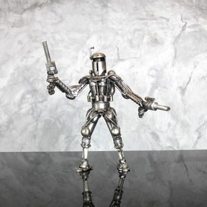 May include: A silver robot figurine made of metal parts, such as nuts, bolts, and screws. The robot is holding a weapon in each hand and is standing on a black surface.