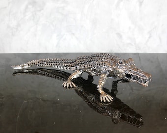 May include: A metal sculpture of a crocodile, made from recycled metal parts. The crocodile is posed in a walking position with its mouth open. The sculpture is silver in color and has a realistic appearance.