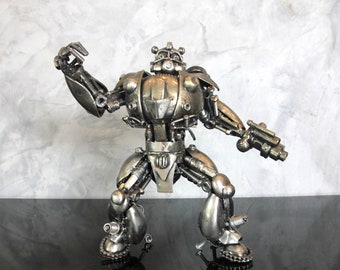 May include: A silver robot sculpture made from recycled metal parts. The robot is standing with its arms raised, one holding a weapon. The sculpture is on a black surface.