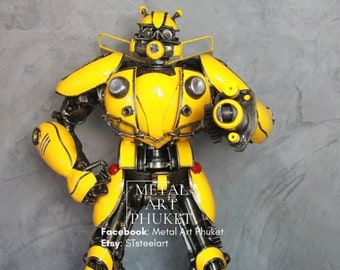May include: A yellow and black robot made of metal, with a detailed design and a gun in its right hand. The robot is standing on a black surface. The text "Metal Art Phuket" is visible on the robot's leg.  Facebook: Metal Art Phuket Etsy: STsteelart