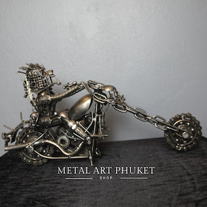 May include: A motorcycle sculpture made entirely of metal parts, including nuts, bolts, and gears. The motorcycle is in a riding position with a rider figure. The sculpture is on a black surface. METAL ART PHUKET SHOP