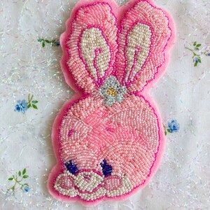 May include: A pink beaded bunny with white ears and a flower on its head. The bunny is sitting on a white background with a floral pattern.