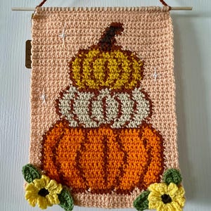 May include: A crocheted wall hanging featuring a stack of three pumpkins. The bottom pumpkin is orange, the middle pumpkin is white, and the top pumpkin is yellow. The pumpkins are decorated with white stitches to resemble the shape of a pumpkin. The wall hanging is decorated with green leaves and yellow flowers.