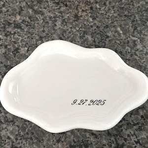 May include: A white, cloud-shaped ceramic dish with a smooth surface. The date "9.27.2025" is printed in black script on the dish. The dish is set against a speckled gray background.