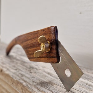 Peut inclure: Gros plan d'un cutter avec un manche en bois et un écrou papillon en laiton. La lame argentée est inclinée. Le manche présente un design incurvé et un grain de bois foncé. Outil de coupe.