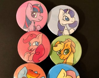Può includere: Sei bottoni rotondi con pony dei cartoni animati in vari colori. Ogni bottone ha un design diverso di pony, tra cui pony rosa, blu, gialli e arancioni. I bottoni sono disposti su una superficie nera.