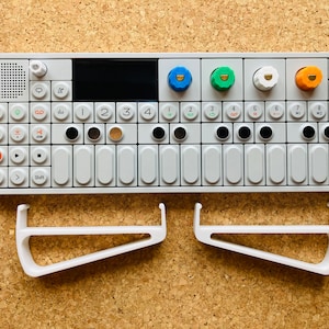 May include: A white electronic music synthesizer with a keyboard, knobs, and buttons. The synthesizer is labeled "OP-1". Two white plastic stands are positioned below the synthesizer.