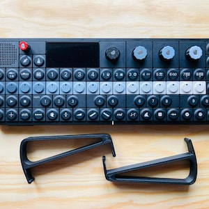 Può includere: Dispositivo nero per la produzione di musica elettronica con layout di tastiera, numerosi pulsanti e manopole. Il dispositivo ha un piccolo schermo e una manopola rossa. Due supporti neri sono sotto. Su una superficie di legno.