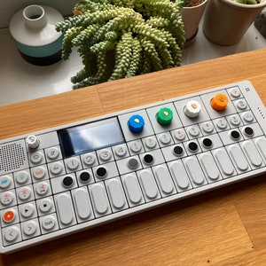 Könnte beinhalten: Ein weißer elektronischer Musiksynthesizer mit einem kleinen Bildschirm, schwarzen Knöpfen und orangefarbenen, grünen und blauen Drehknöpfen. Der Synthesizer ist mit "OP-1" beschriftet.