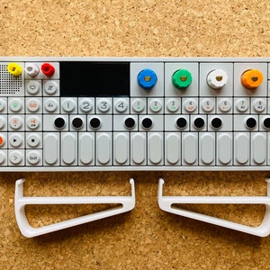 Peut inclure: Un synthétiseur blanc avec un clavier noir et des boutons colorés. Le synthétiseur est étiqueté "OP-1" et possède une variété de boutons et de commandes. Le synthétiseur est posé sur une surface en liège.