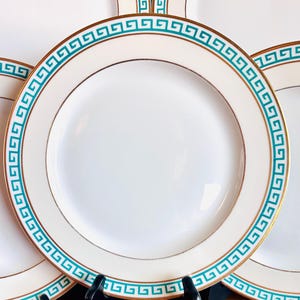May include: White ceramic plates with a teal and gold Greek key border design. The plates have a gold rim and are displayed on black stands against a wooden surface. The plates are a classic tableware item.