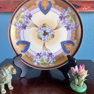 May include: A decorative plate with a gold rim and a floral design featuring purple flowers and green leaves. The plate is supported by a black stand. A small bronze dog figurine and a miniature tulip arrangement are also present.