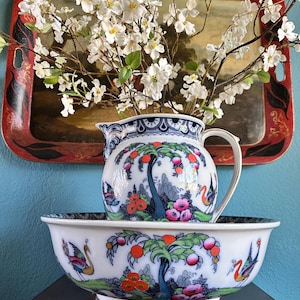 May include: A decorative porcelain pitcher and bowl set with a colorful, detailed design of trees, flowers, and birds. The pitcher is filled with white flowers. The set is placed on a dark wooden surface against a blue wall.