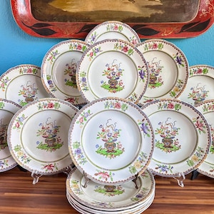 Peut inclure: Ensemble d'assiettes en céramique blanche avec un motif floral. Chaque assiette présente un arrangement floral central dans un vase, entouré d'une bordure de fleurs colorées. Les assiettes sont disposées sur une surface en bois.