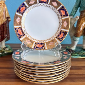Op de afbeelding: Een stapel fijn porseleinen borden met een witte kern en een decoratieve rand met oranje bloemmotieven, marineblauwe accenten en gouden rand. De borden worden gepresenteerd op een houten oppervlak.