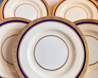 May include: A set of five white ceramic plates featuring a wide cobalt blue border and intricate gold filigree patterns. Each plate has a central white circular area surrounded by a gold ring, with a decorative gold-trimmed edge.
