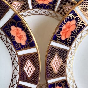 Puede incluir: Primer plano de varios platos de porcelana blanca con diseños intrincados. Los platos presentan un borde azul marino con detalles dorados, secciones rectangulares blancas y motivos florales naranjas. Los bordes son dorados.