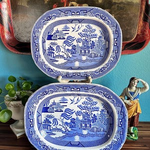 May include: Two blue and white oval platters with a Willow pattern design. The platters are displayed on white stands. The design features a bridge, pagoda, and birds. Classic tableware for serving food.