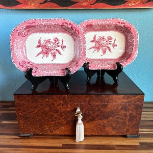 May include: Two pink and white oval serving dishes with floral designs are displayed on a wooden box with a white tassel. The dishes are on black stands.