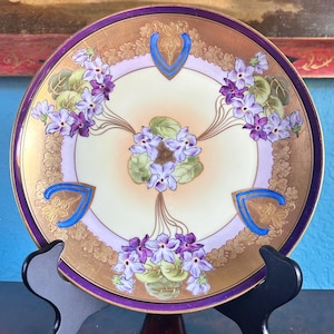 May include: A vintage decorative plate with a gold rim and floral pattern. The plate features purple flowers, green leaves, and blue accents. It is displayed on a black stand, highlighting its intricate design and antique appeal.