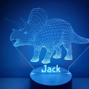 K&ouml;nnte beinhalten: Ein blaues, 3D-gedrucktes Dinosaurier-Nachtlicht mit dem Namen "Jack" darunter. Das Licht befindet sich auf einem wei&szlig;en Sockel mit einem Ein-/Ausschalter.