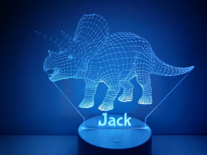 K&ouml;nnte beinhalten: Ein blaues, 3D-gedrucktes Dinosaurier-Nachtlicht mit dem Namen "Jack" darunter. Das Licht befindet sich auf einem wei&szlig;en Sockel mit einem Ein-/Ausschalter.