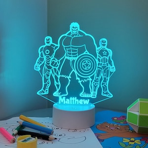 May include: A blue LED night light showing the Hulk, Captain America, and Iron Man. The base is white, and the name "Matthew" is displayed. The light sits on a table with crayons and colouring pages.