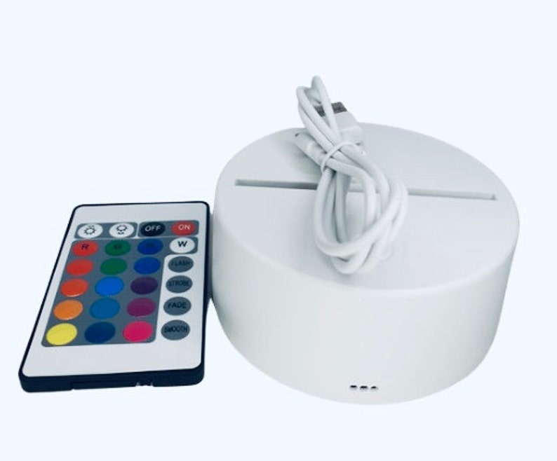 K&ouml;nnte beinhalten: Eine wei&szlig;e runde LED-Lichtbasis mit einem USB-Kabel und einer Fernbedienung. Die Fernbedienung hat Tasten zum Ein- und Ausschalten, zur Farbauswahl und f&uuml;r Lichteffekte.