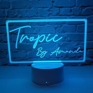 May include: A white acrylic sign with a blue light behind it. The sign says "Tropic By Amanda" in a cursive font.