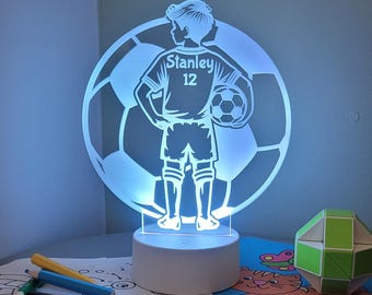 Puede incluir: Una lámpara de noche LED brillante con un diseño de jugador de fútbol. La luz tiene la forma de un balón de fútbol con un jugador sosteniendo un balón. La camiseta del jugador tiene el nombre "Stanley" y el número "12".