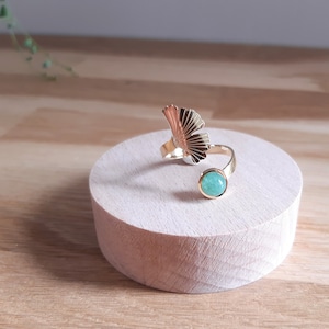 May include: A gold-colored ring with a fan-shaped design and a round, light blue gemstone. The ring is displayed on a small, round wooden stand, showcasing its intricate details and elegant style.