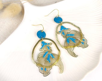 May include: Gold-toned fish-shaped earrings with blue accents. The earrings feature a detailed fish design within a circular frame, with a blue circular stud at the top. The fish have blue details and are suspended from gold-colored ear wires.