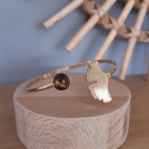 May include: Gold-toned bracelet with a ginkgo leaf design and a round, brown stone. The bracelet is displayed on a light wooden stand. The ginkgo leaf is on one end, and the stone is on the other. The background is a light blue wall.
