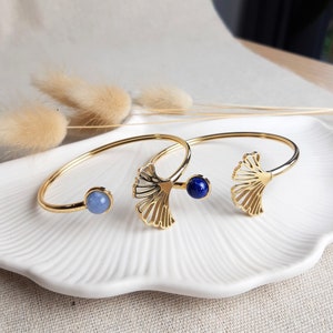 Op de afbeelding: Twee gouden armbanden met ginkgo-bladeren. De ene armband heeft een lichtblauwe steen, terwijl de andere een donkerblauwe steen heeft.