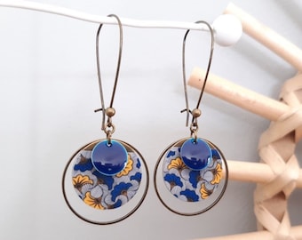 Può includere: Un paio di orecchini pendenti con un motivo floreale blu e giallo su sfondo bianco. Gli orecchini hanno un cerchio in bronzo e un cerchio blu al centro.