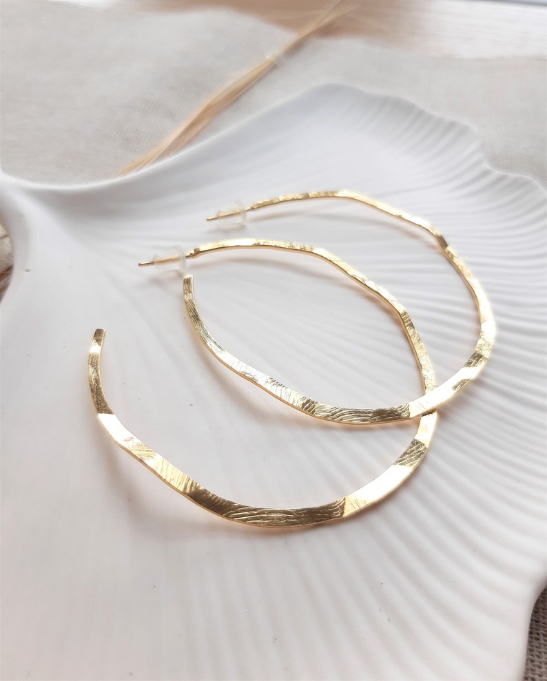 May include: A pair of gold hoop earrings with a textured finish. The hoops are large and have a slightly oval shape.