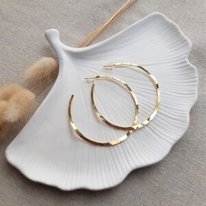 May include: A pair of gold hoop earrings with a hammered texture. The earrings are displayed on a white ceramic dish.