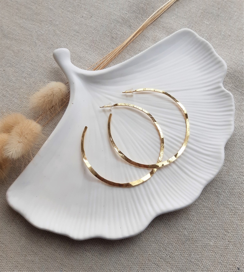 May include: A pair of gold hoop earrings with a hammered texture. The earrings are displayed on a white ceramic dish.