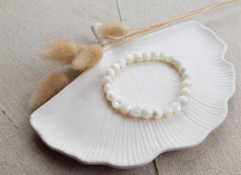 K&ouml;nnte beinhalten: Ein wei&szlig;es Armband aus Muschelkernperlen auf einem wei&szlig;en Keramikteller mit F&auml;cherform. Das Armband besteht aus runden, wei&szlig;en Perlen mit Perlmutt-Finish.
