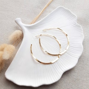 May include: Two gold hoop earrings with a hammered texture, displayed on a white ceramic dish shaped like a leaf.