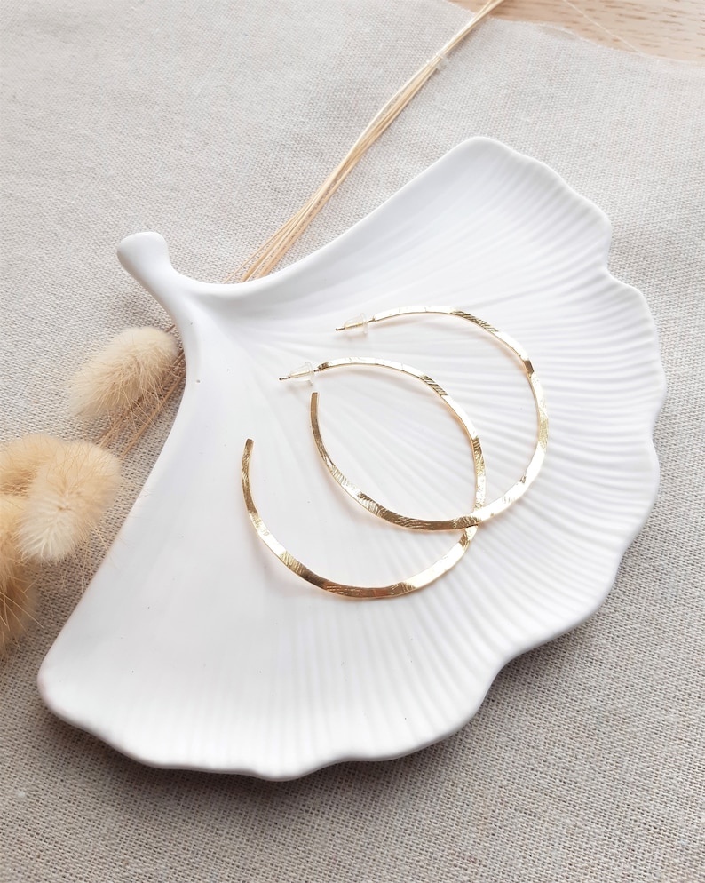 May include: Two gold hoop earrings with a hammered texture, displayed on a white ceramic dish shaped like a leaf.
