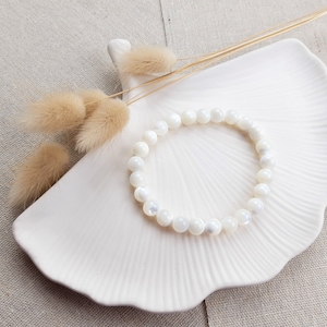 K&ouml;nnte beinhalten: Ein Armband aus wei&szlig;en Muschelkernperlen auf einer wei&szlig;en Keramikplatte mit getrockneten Blumen.